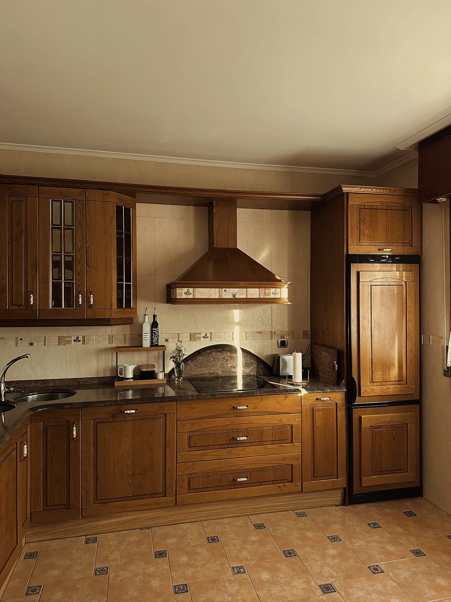 Une cuisine spacieuse avec des meubles en bois, un réfrigérateur assorti, une cuisinière encastrée, un four et une hotte aspirante. Le plan de travail accueille une bouilloire, un grille-pain et quelques bouteilles. Le carrelage au sol et les murs beiges complètent ce style classique et chaleureux.