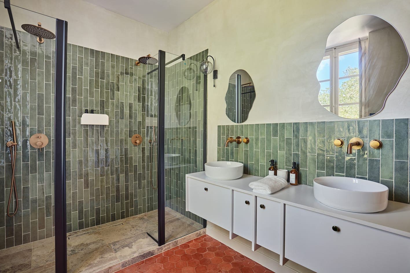 Salle de bain moderne avec carrelage vertical vert dans la douche, deux vasques rondes sur une vanité blanche, des luminaires dorés, deux miroirs de forme irrégulière et une fenêtre laissant entrer la lumière naturelle.