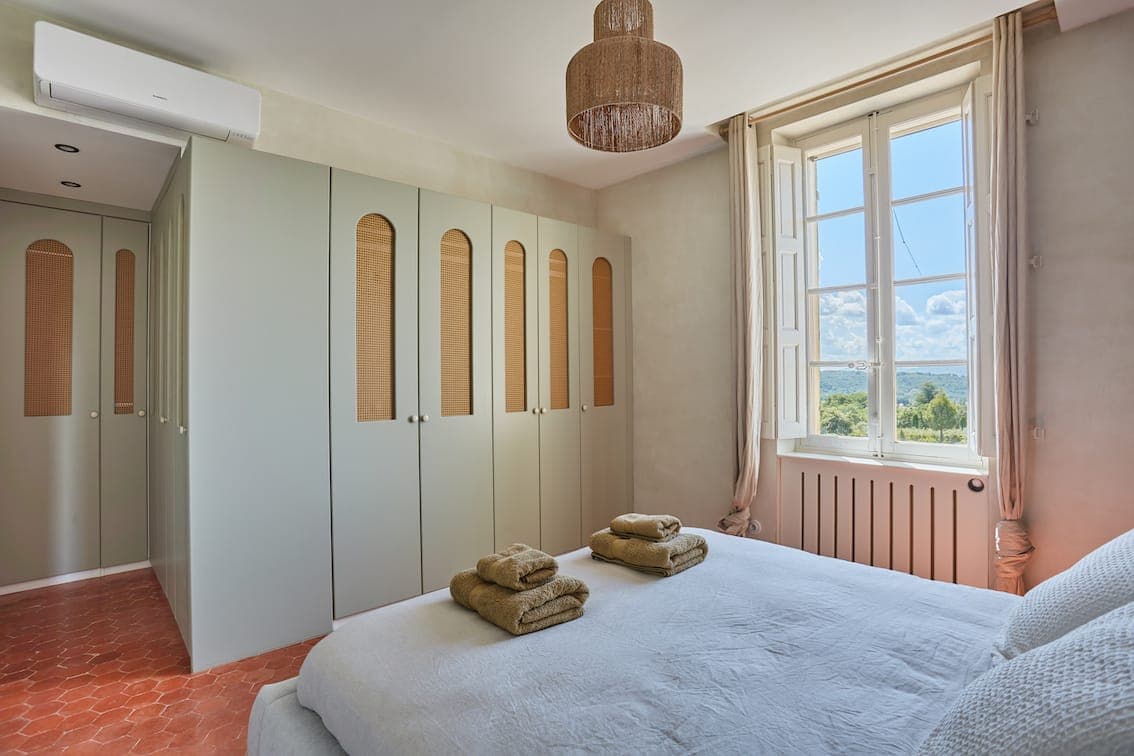 Une chambre lumineuse et minimaliste avec un lit soigneusement fait, deux ensembles de serviettes pliées, des armoires vert clair, une grande fenêtre avec une vue panoramique, de la lumière naturelle et un sol en carrelage en terre cuite.