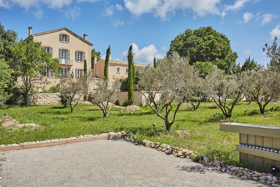 Une grande maison beige de trois étages avec des volets bleus se trouve derrière un jardin avec des oliviers, des murs en pierre et un chemin de gravier sous un ciel partiellement nuageux.