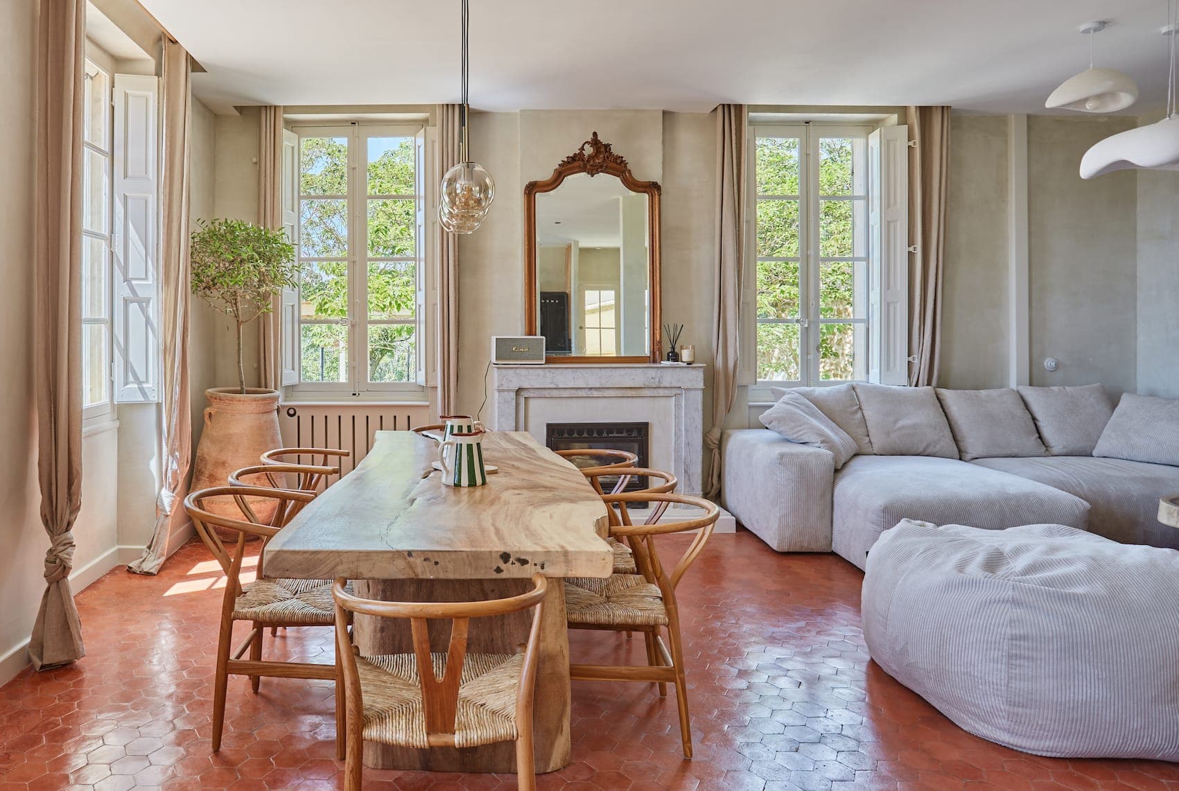 Salon et salle à manger lumineux avec de grandes fenêtres, une table à manger en bois avec des chaises, un canapé sectionnel gris, un pouf, un arbre en pot, une cheminée avec un miroir au-dessus et un sol en carrelage en terre cuite.