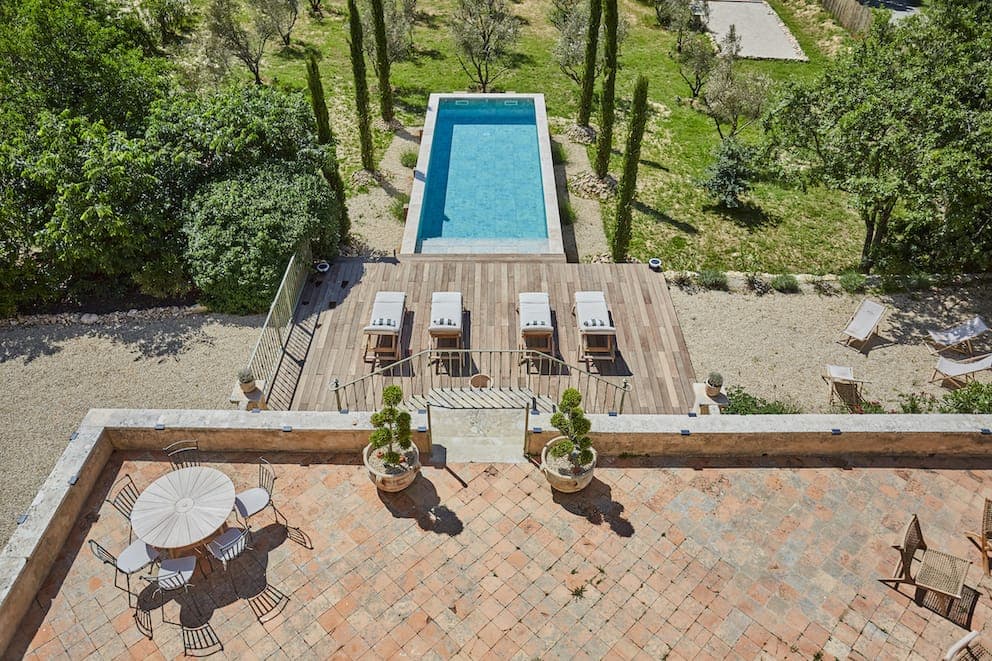 Vue aérienne d'une cour arrière avec une piscine rectangulaire, quatre chaises longues sur une terrasse en bois et un patio carrelé avec une table ronde et des chaises, entouré de verdure luxuriante et d'arbres.