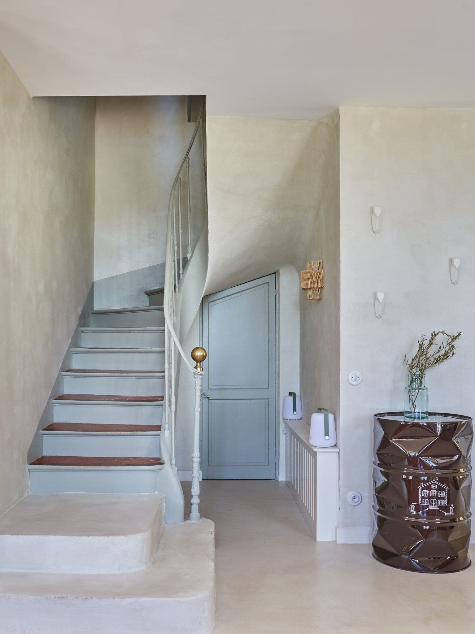 Un couloir minimaliste avec un escalier en colimaçon clair, une porte bleu clair sous l'escalier et un tonneau décoratif marron foncé avec des branches dans un vase à proximité. Les murs et le sol sont clairs et neutres.