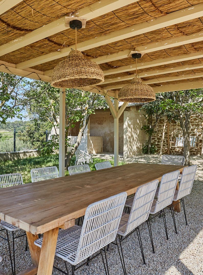 Une longue table à manger d'extérieur en bois avec des chaises blanches tressées se trouve sous une pergola avec un toit en roseau et deux suspensions en osier, entourée d'arbres et de verdure sur un patio en gravier.