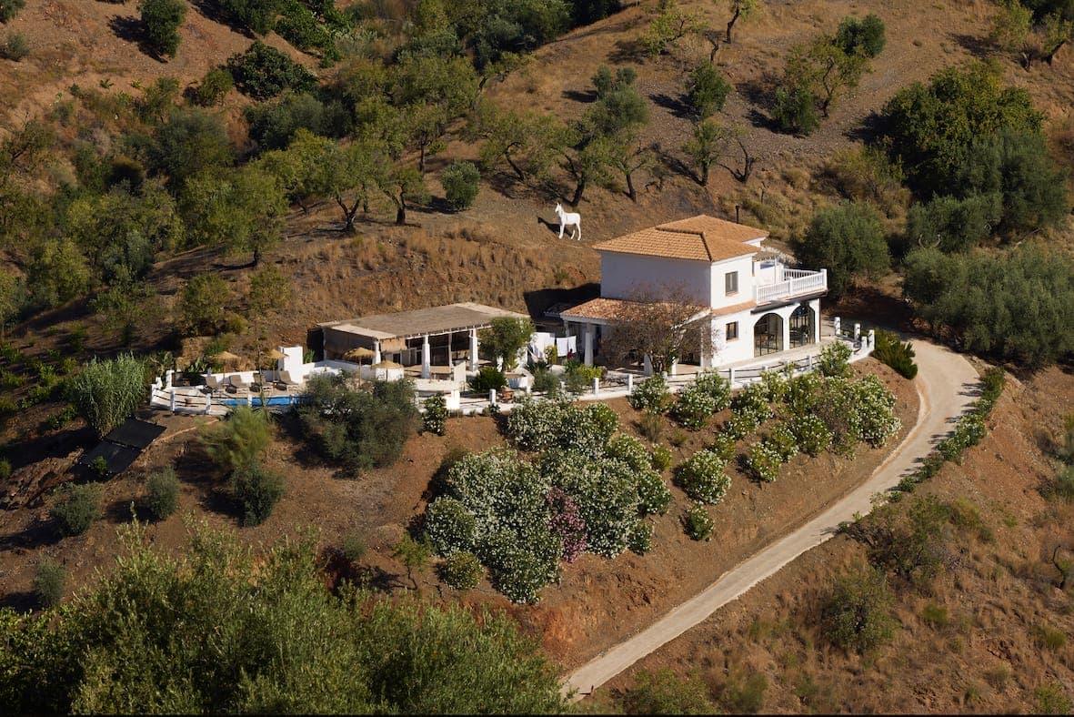 A white house with a terracotta roof stands on a hillside, surrounded by greenery and trees. A winding dirt road leads to the house. A large metal horse sculpture can be seen on the hill above.