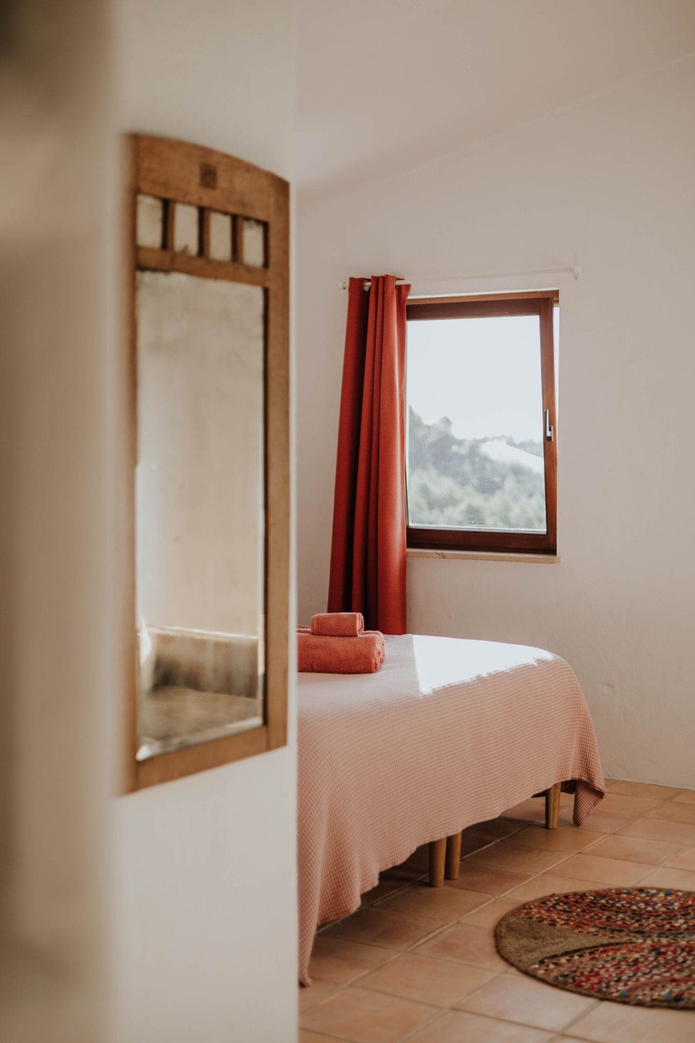 A cozy bedroom with a bed covered in a light pink blanket, topped with neatly folded pink towels. A window with red curtains lets in natural light. A mirror and a round rug are visible in the warm, minimalist room.