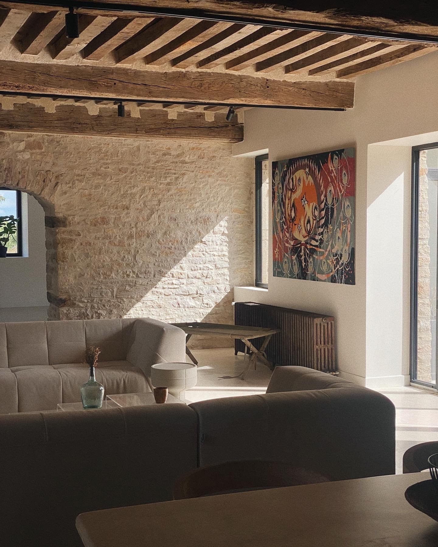 Sunny living room with beige corner sofa, wooden beams and stone wall. A colorful abstract painting hangs above a radiator. Large windows let in natural light, casting shadows on the floor, and a plant is visible outside.