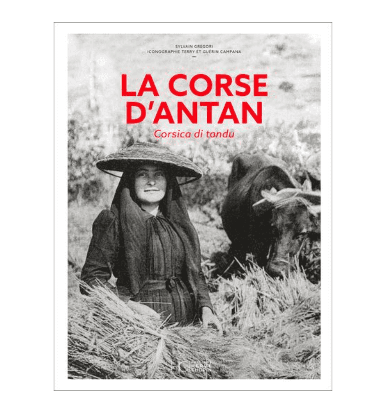 Photographie vintage en noir et blanc d'une femme souriante, vêtue d'un costume traditionnel et coiffée d'un chapeau à larges bords, travaillant dans un champ, une vache derrière elle. Le texte « La Corse d'Antan » et « Corsica di tandù » est écrit en lettres rouges.