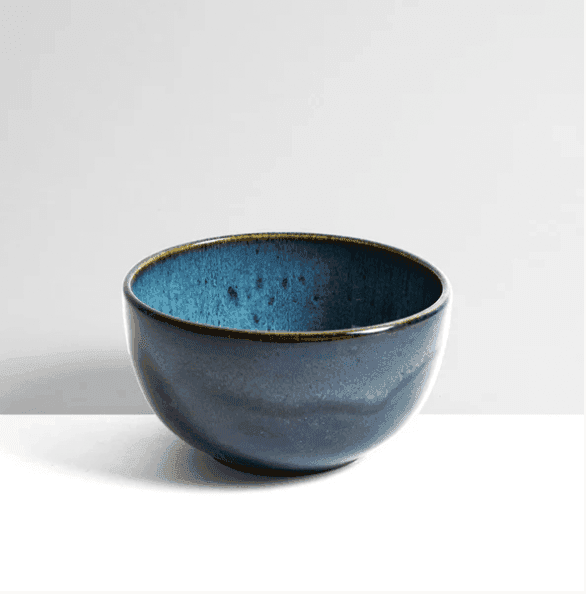 Un bol en céramique à l'extérieur émaillé bleu foncé brillant et à l'intérieur texturé bleu plus clair. Ce bol présente un subtil motif moucheté et repose sur une surface neutre sur un fond gris clair uni.