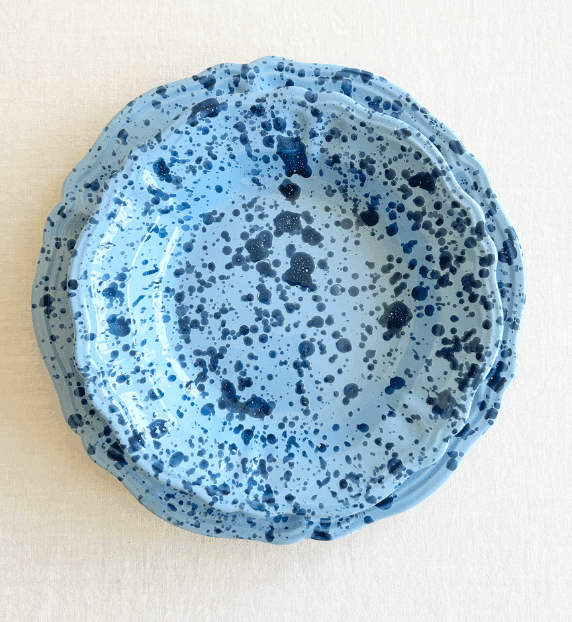 Deux assiettes rondes en céramique bleu clair avec des bords festonnés et des motifs d'éclaboussures bleu foncé, empilées sur une surface texturée blanche.