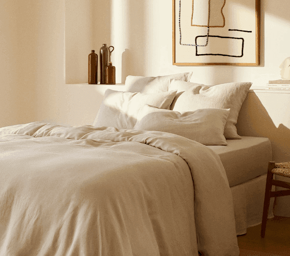 Une chambre douillette avec un lit impeccablement fait, des draps beige clair et de nombreux oreillers. La pièce bénéficie d'un éclairage chaleureux et d'une décoration minimaliste, avec notamment une œuvre d'art abstraite encadrée au mur et des vases décoratifs sur une étagère.