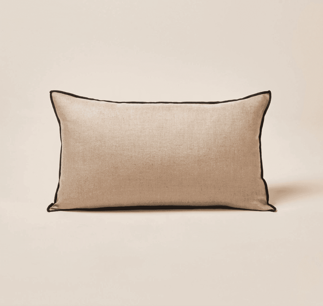 Un coussin rectangulaire beige avec un passepoil noir le long des bords, posé sur un fond neutre.