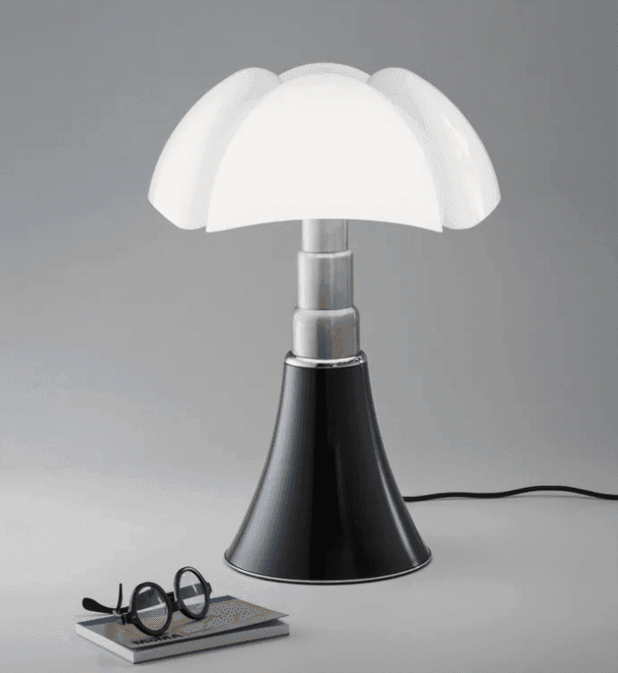 Une lampe de table moderne, dotée d'un abat-jour blanc en forme de pétale et d'une base noire évasée, est posée sur une surface plane, à côté d'une paire de lunettes rondes noires et d'un carnet fermé. Le fond est gris clair uni.