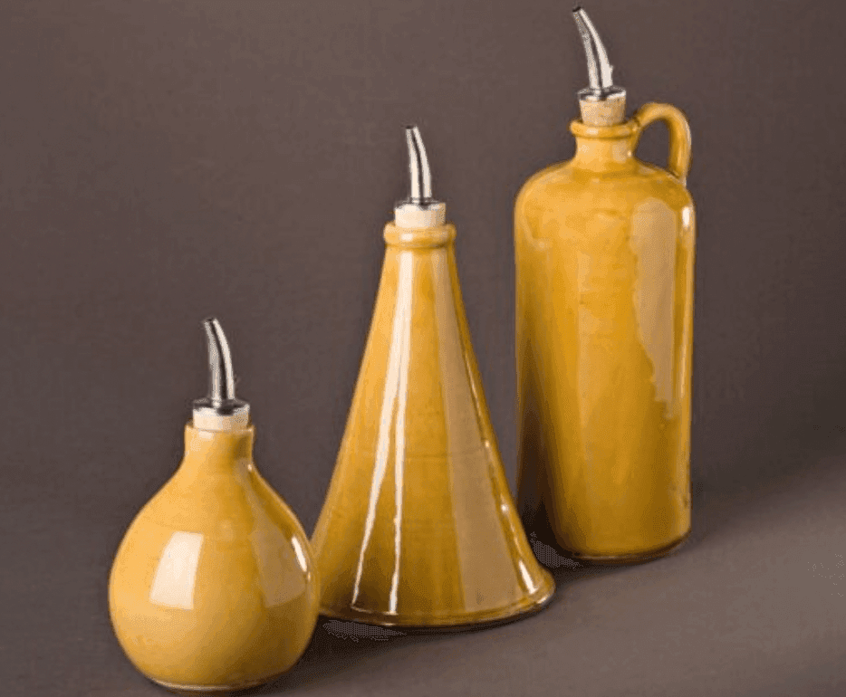 Trois huiliers en céramique de formes et de tailles variées, dotés de becs verseurs, de couleur jaune moutarde, sont disposés sur un fond gris foncé.