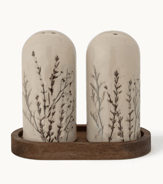 Deux shakers en céramique sur un plateau en bois, décorés de délicats motifs de plantes et de feuilles, sur un fond blanc uni.