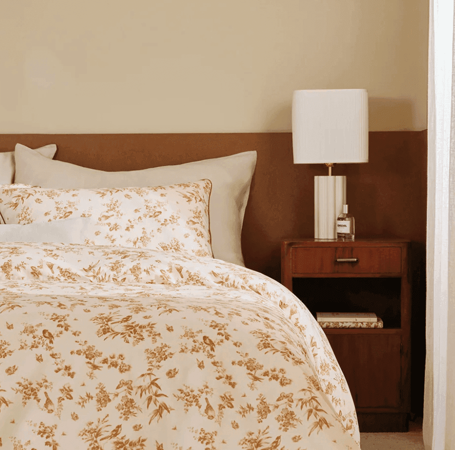 Une chambre douillette avec un lit orné de parures de lit à fleurs blanches et dorées, des oreillers beiges et crème et une tête de lit capitonnée marron. Une table de chevet en bois accueille une lampe blanche, un livre et une bouteille en verre. La lumière filtre par la droite.