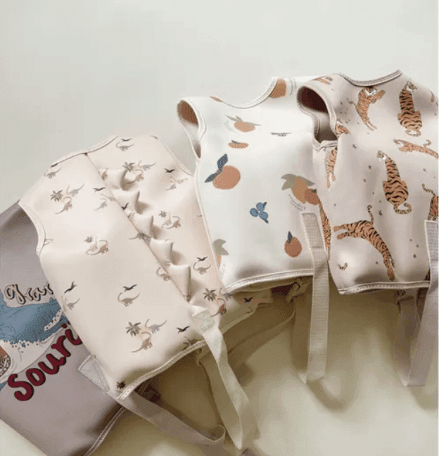 Trois gilets de natation pour enfants sont exposés sur une surface blanche. Chaque gilet arbore un motif différent : dinosaures, fruits et tigres. Un sac gris partiellement imprimé se trouve à côté des gilets.