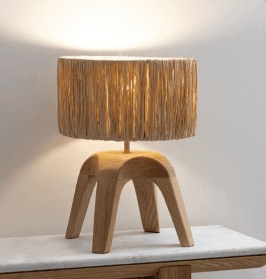 Une lampe de table moderne avec un abat-jour cylindrique en rotin tressé et une base en bois unique en forme d'arche, placée sur une surface en marbre de couleur claire contre un mur blanc uni.