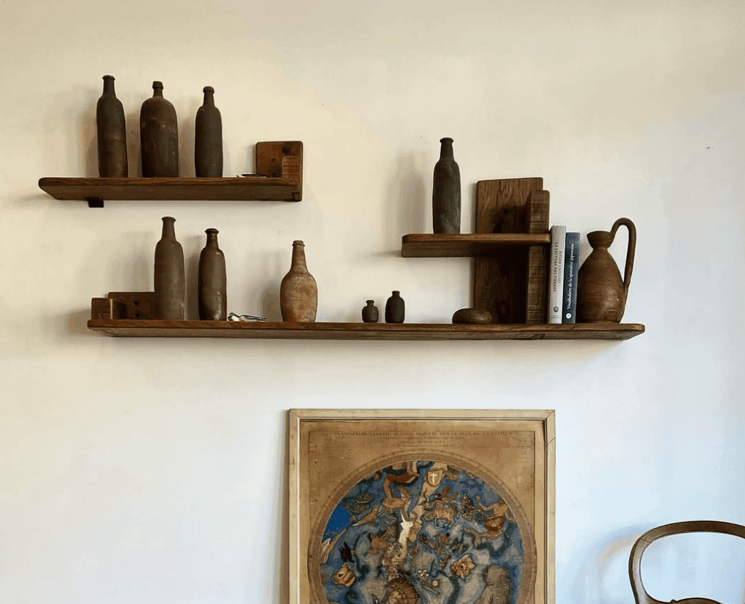Étagères en bois sur un mur blanc présentant divers objets en poterie, dont des bouteilles et des cruches. En dessous, une carte encadrée de style vintage. Une vue partielle d'une chaise en bois est visible à droite.