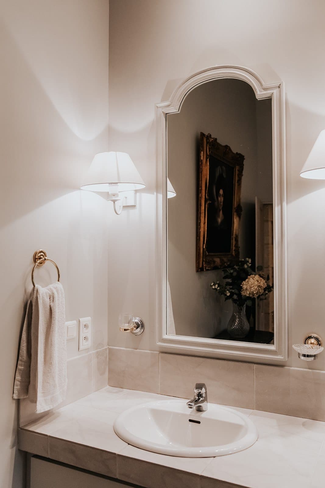 Une salle de bain avec un lavabo blanc, un plan de travail en marbre, un anneau porte-serviettes avec une serviette blanche, des appliques murales, un grand miroir encadré et un portrait encadré qui se reflète dans le miroir. Un vase rempli de fleurs est également visible dans le reflet.