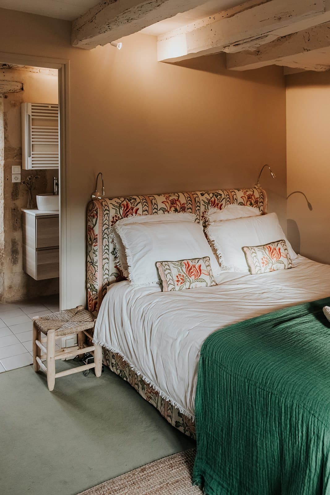 Chambre confortable avec un lit tapissé de fleurs, une parure de lit blanche et une couverture verte. Deux petits coussins à motifs floraux rouges sont posés sur le lit. Un tabouret en bois se trouve à côté du lit, avec une salle de bain visible en arrière-plan.