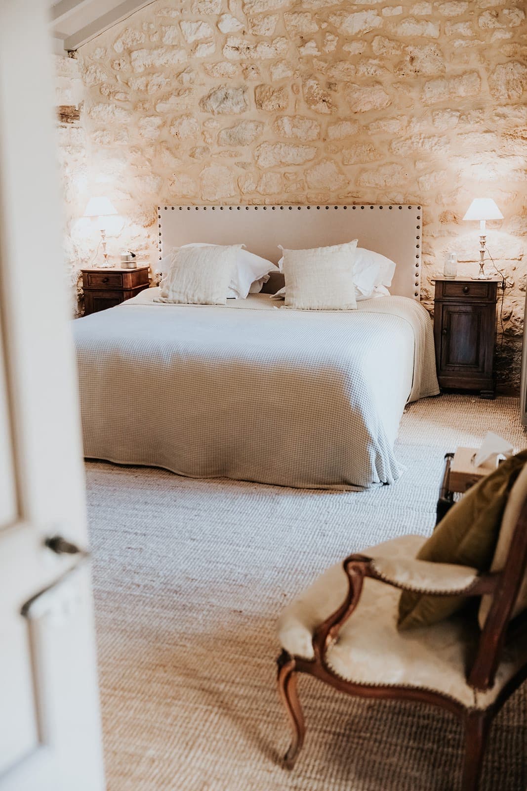 Chambre cosy avec un grand lit, une literie aux tons neutres, deux tables de chevet avec lampes, un mur en pierres apparentes et un fauteuil rembourré au premier plan. Un éclairage doux et chaleureux crée une atmosphère paisible.