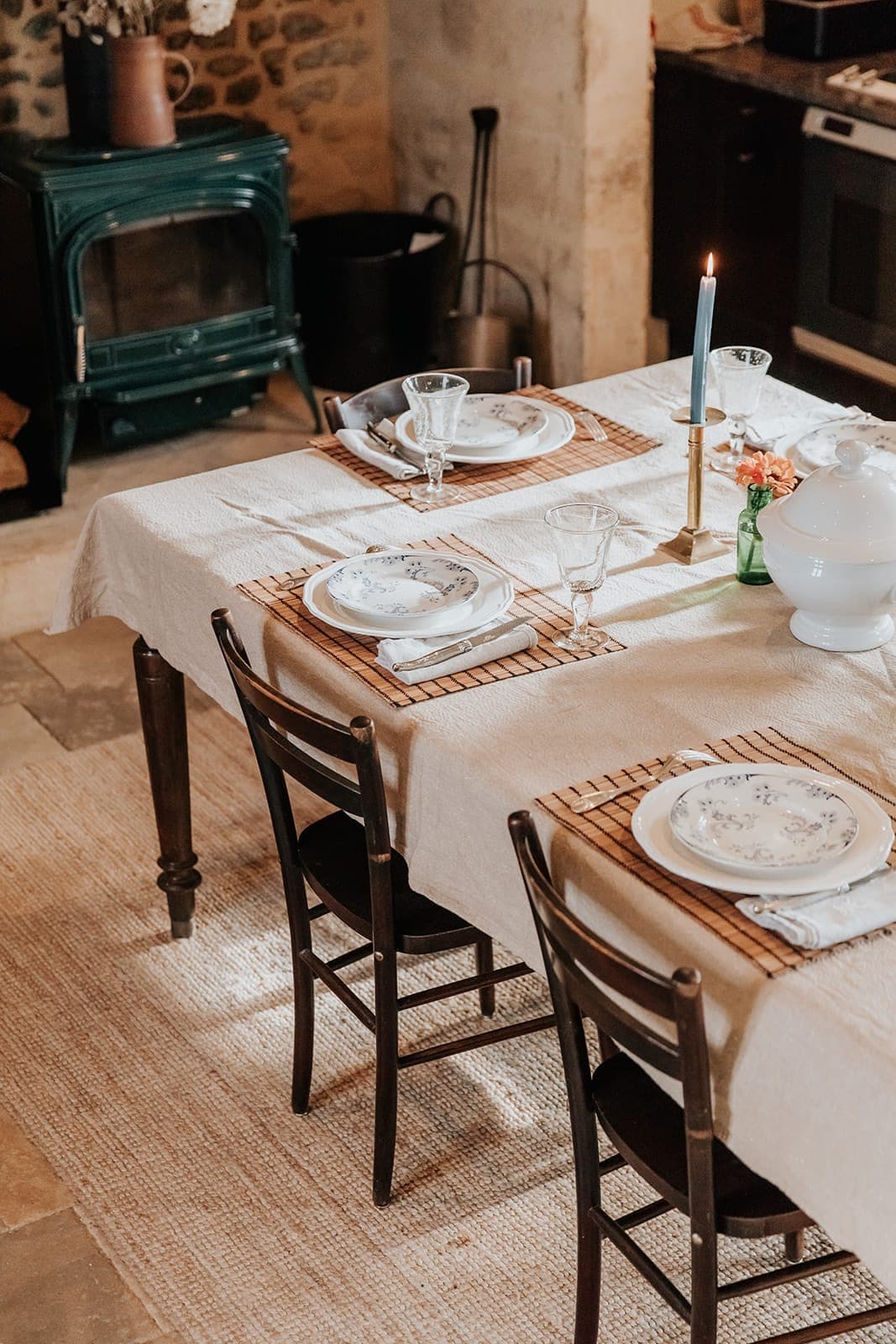 Une table à manger rustique dressée pour quatre personnes avec des assiettes fleuries, de la verrerie et une seule bougie bleue allumée, placée sur une nappe beige dans une cuisine chaleureuse avec des murs en pierre et un poêle à bois vert.