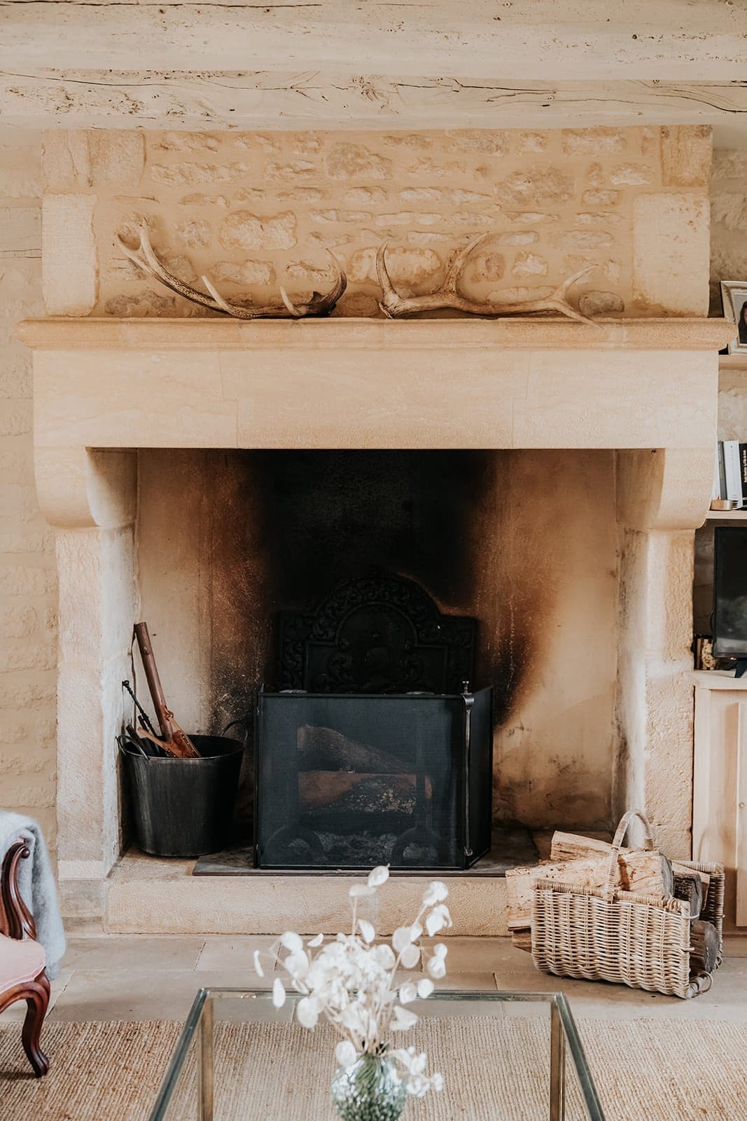 Une cheminée rustique en pierre avec des bois sur le manteau, un pare-feu en métal, des bûches et des outils de cheminée à gauche et un panier en osier avec plus de bûches à droite, dans un cadre de salon chaleureux.