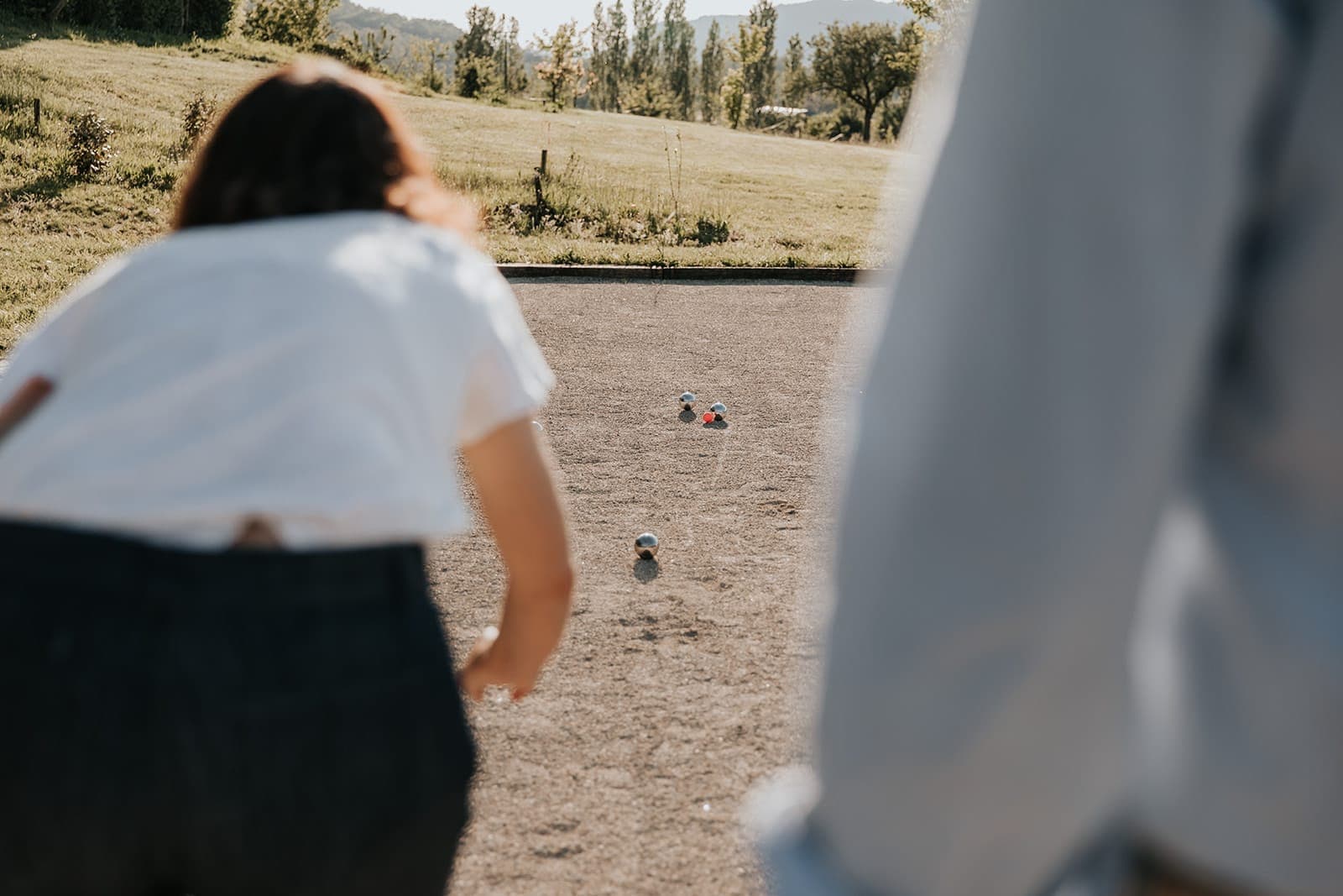 Deux personnes jouent à la pétanque en plein air par une journée ensoleillée. Une personne est au premier plan, floue, s'apprêtant à lancer une boule vers plusieurs boules colorées sur un terrain en gravier. L'herbe verte et les arbres sont à l'arrière-plan.
