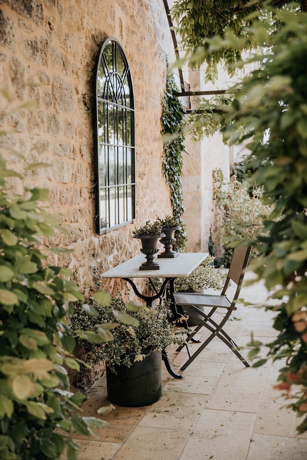 Une petite table avec deux plantes en pot et une chaise pliante se trouve à côté d'un mur de pierre et d'une fenêtre cintrée, entourée d'un feuillage vert luxuriant dans un cadre de jardin extérieur paisible.
