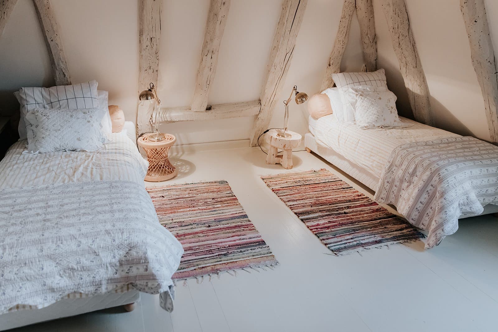 Une chambre confortable avec deux lits simples, une literie blanche à motifs, des poutres en bois rustiques, deux tapis à rayures colorées et de petites tables de chevet avec lampes. L'atmosphère y est chaleureuse et accueillante.