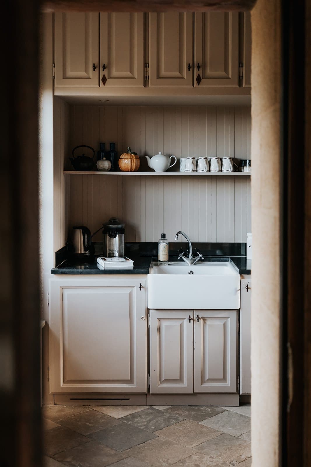 Un coin cuisine confortable avec des armoires crème, un évier de ferme, des étagères ouvertes contenant de la verrerie, une théière, des tasses et une citrouille, ainsi qu'une cafetière et du savon près de l'évier, vus à travers une porte.