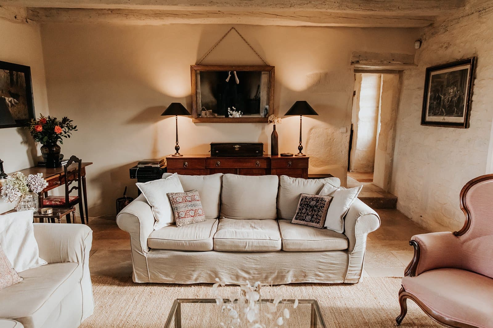Un salon chaleureux avec un canapé beige, des coussins à motifs, une table basse en verre et un fauteuil. Derrière le canapé se trouve un buffet en bois avec des lampes, des fleurs et un miroir encadré au mur.