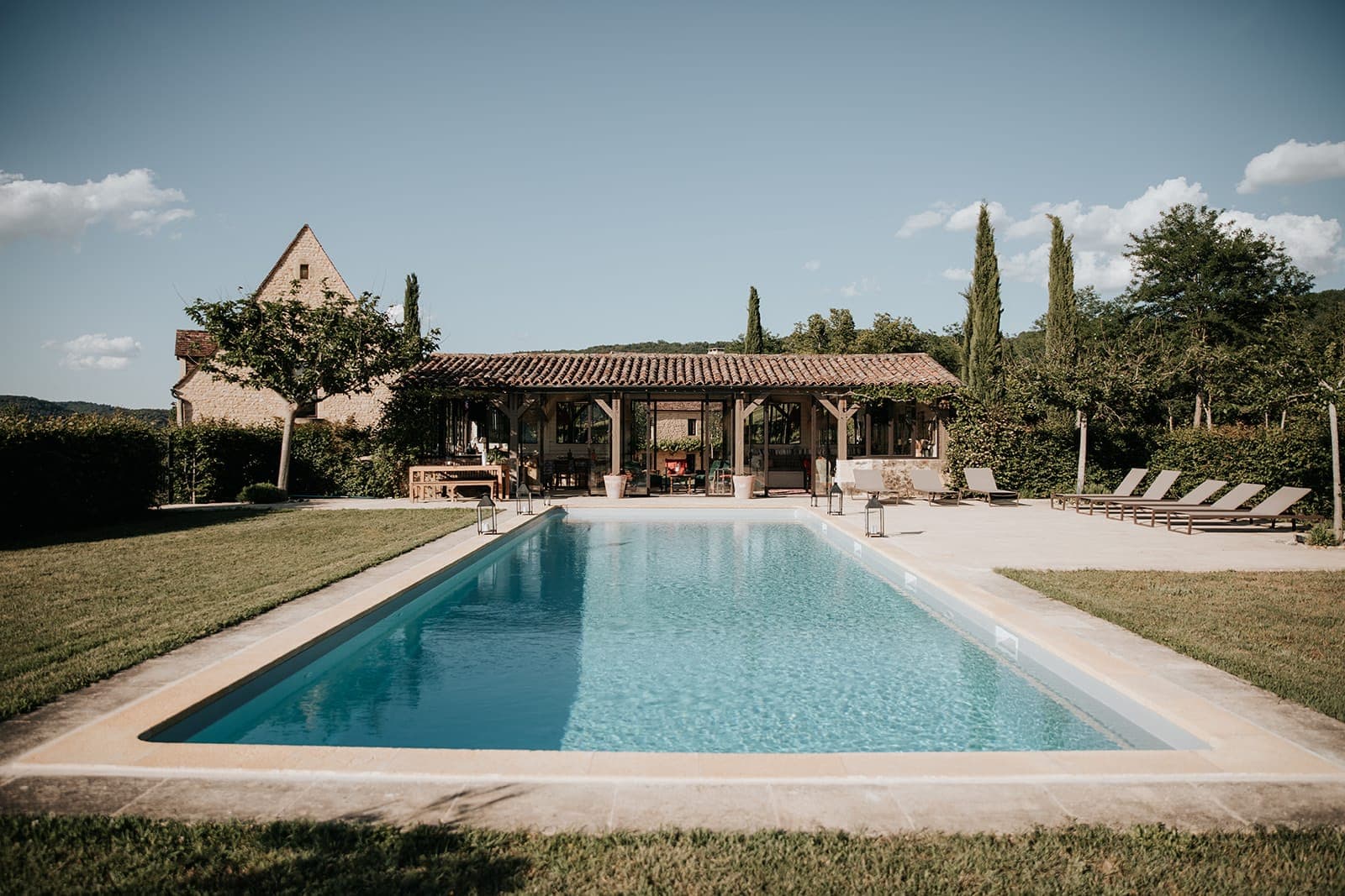Piscine extérieure rectangulaire aux eaux bleues claires, entourée d'un patio et de chaises longues, avec une maison rustique et des arbres en arrière-plan sous un ciel clair.