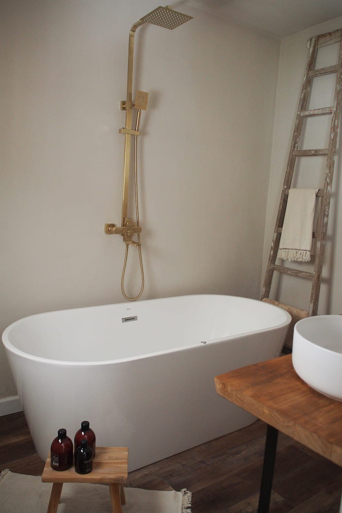 A modern bathroom with a white freestanding bathtub, gold shower faucet, wooden stool with bottles, rustic ladder with towel and wooden countertop with white sink.