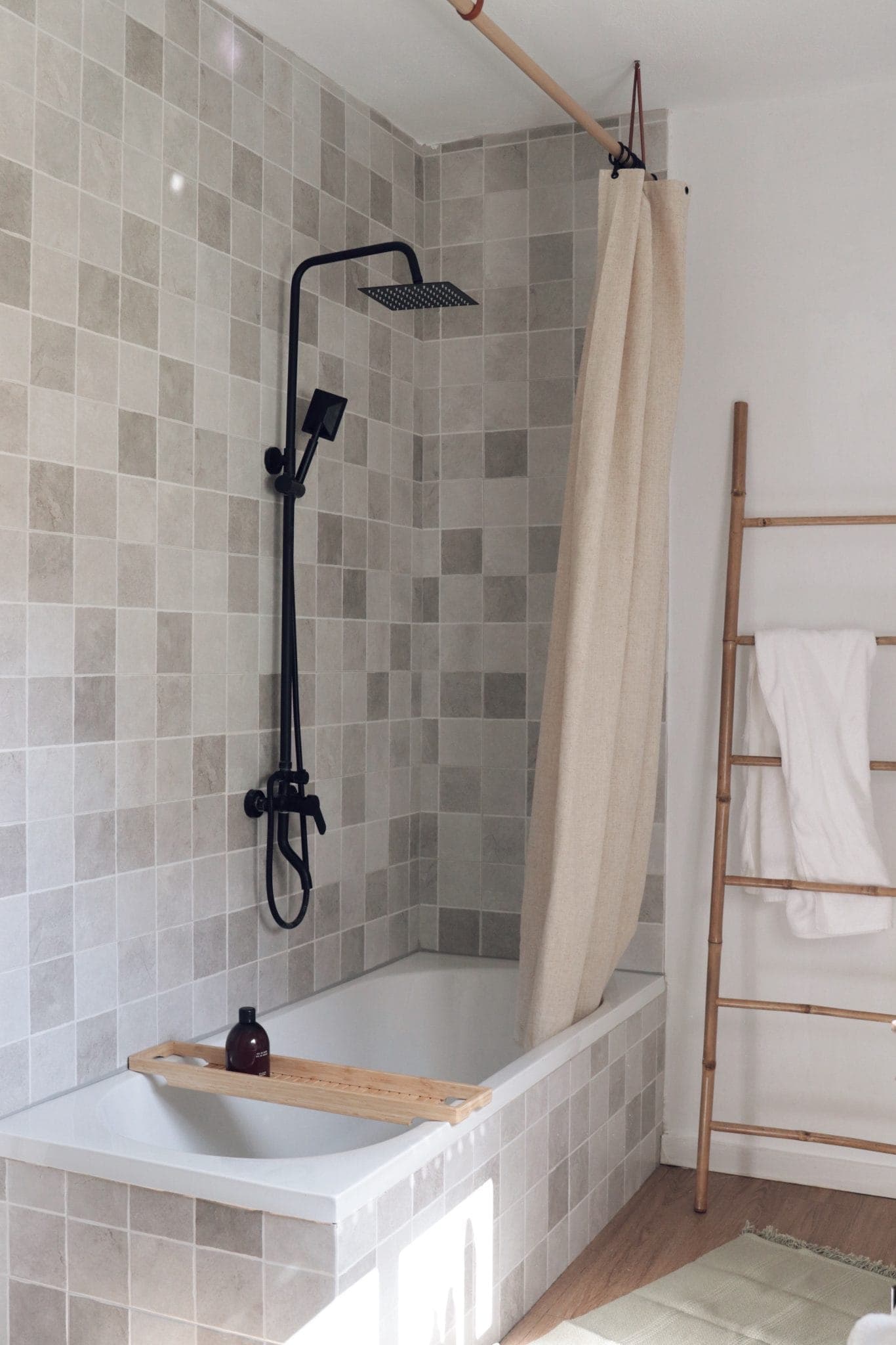 A modern bathroom with light gray square tiles, a white bathtub with a black shower head, a beige shower curtain, a wooden bath tray holding a bottle and a bamboo ladder holding a white towel.