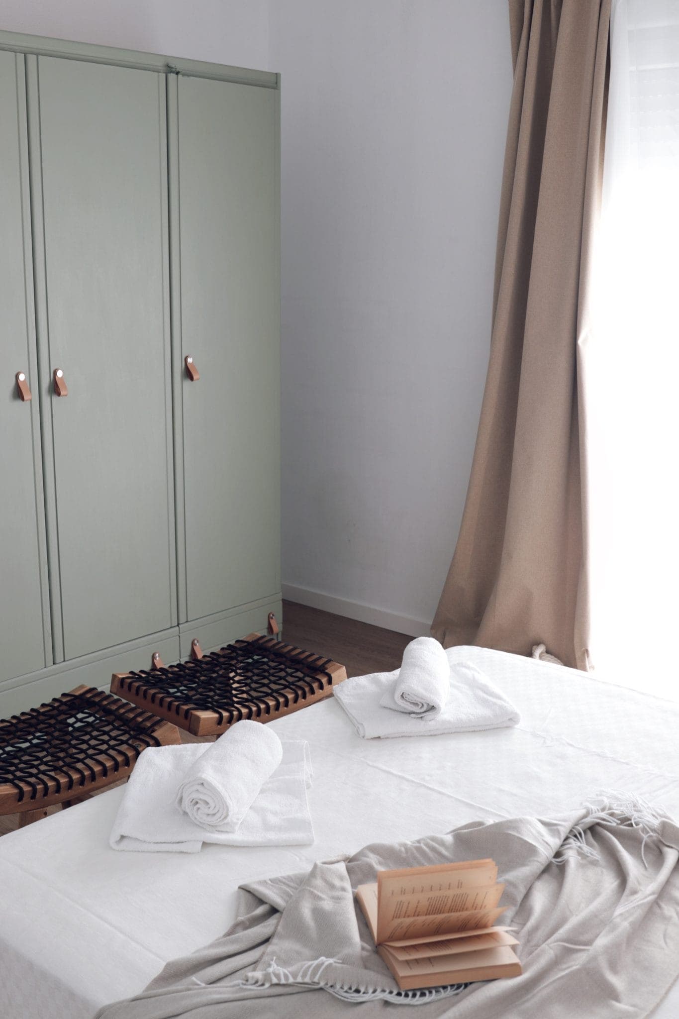 A tidy, minimalist room with a light-green wardrobe, two white towels rolled up on the bed, beige curtains and an open book on a blanket. Two wooden massage mats lie on the floor next to the wardrobe.