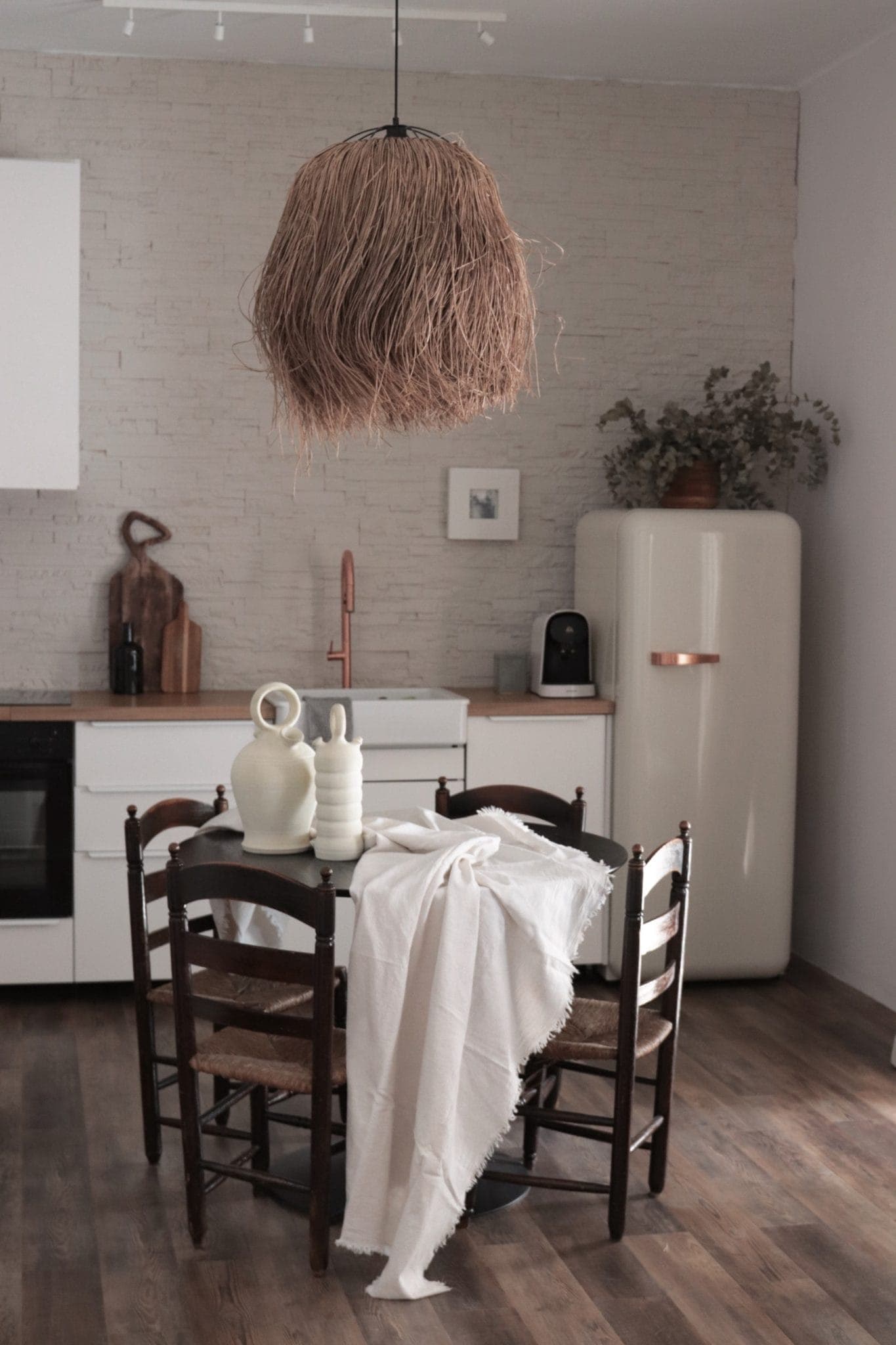 A warm kitchen with parquet flooring, a round table with a white tablecloth, dark wood chairs, white ceramic vases, a retro fridge and a large brown textured pendant hanging above.