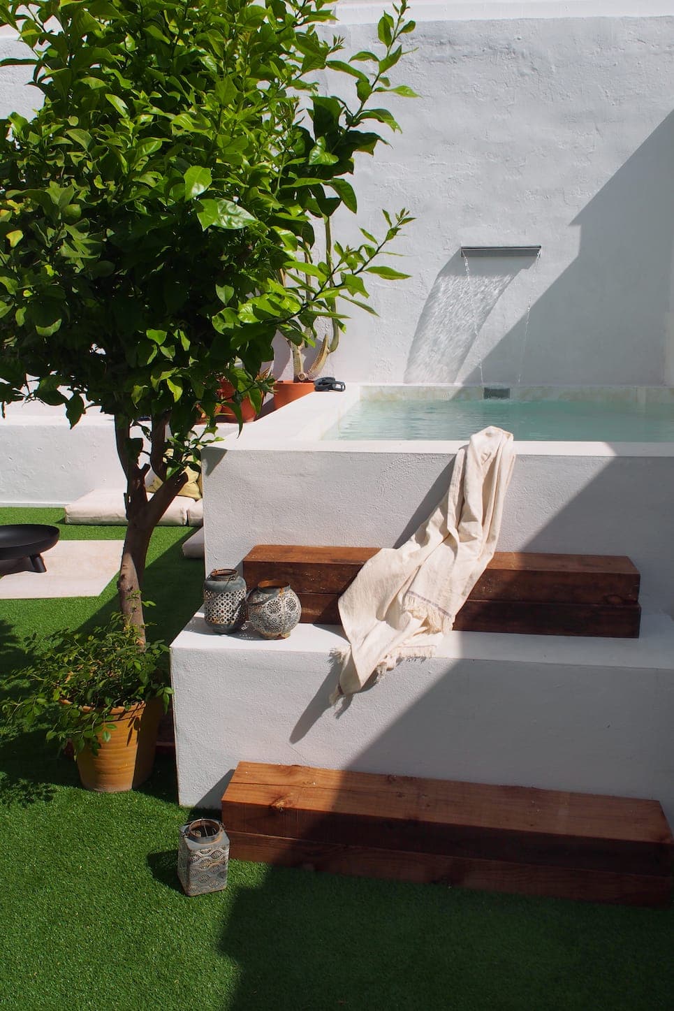 A small white outdoor pool with water flowing from a spout, wooden steps, a cream-colored towel draped over the edge, potted plants, a tree and decorative lanterns on artificial grass in the sun.