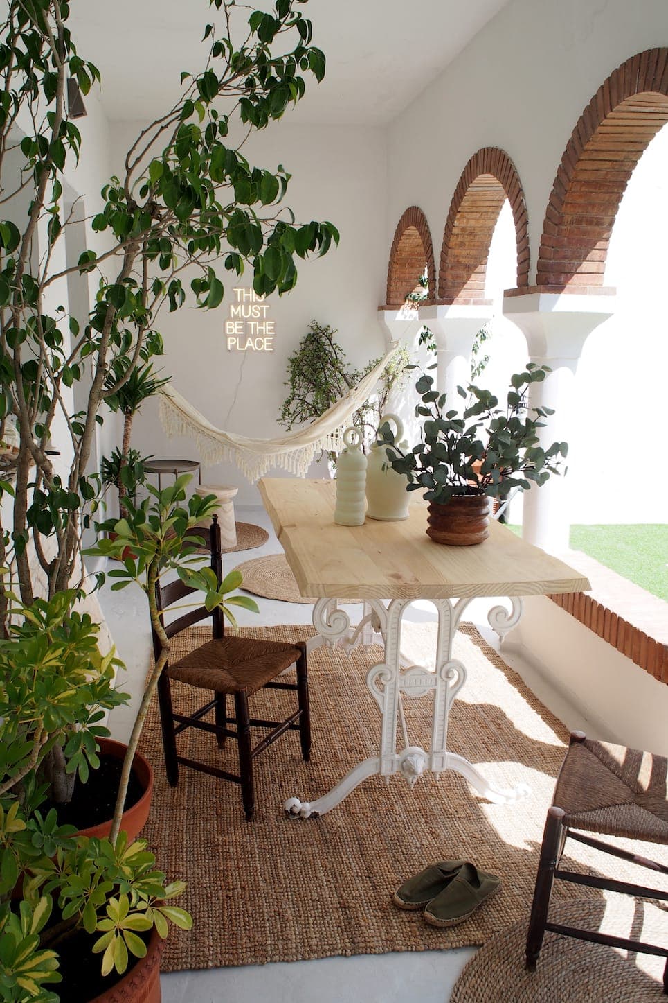 A sunny patio with arched brick columns, a wooden table with chairs, potted plants, a woven rug, a hammock and a neon sign reading “THIS MUST BE THE PLACE” on a white wall.