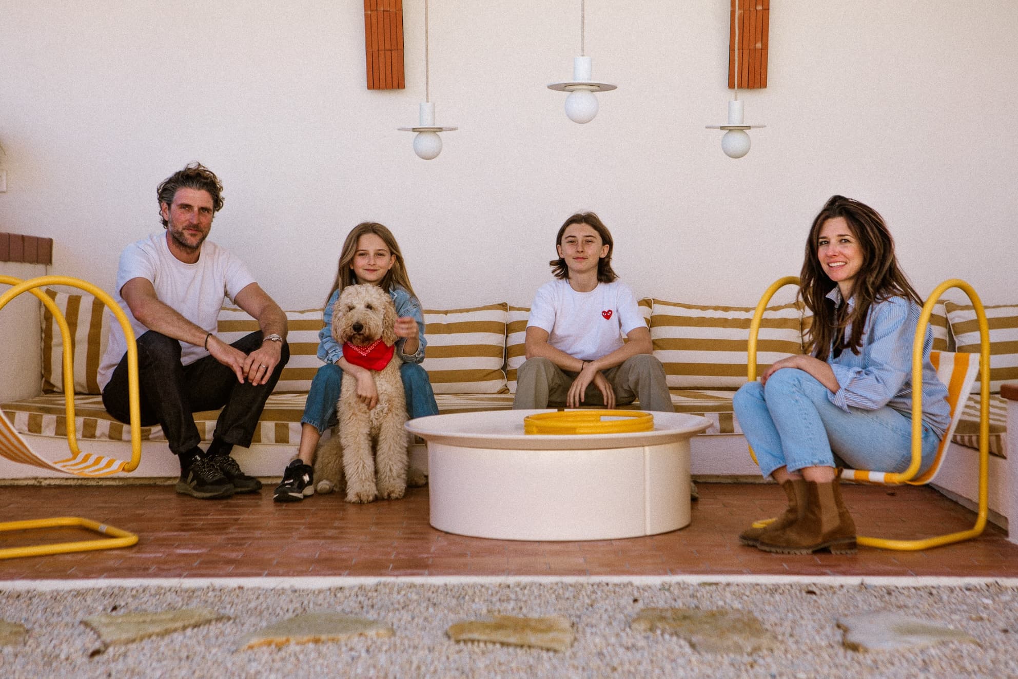 Une famille de quatre personnes est assise sur des bancs rayés en plein air, accompagnée d'un chien tout doux coiffé d'un bandana rouge. Sur la terrasse de Maisons Dominos Audrey Colombani, des chaises jaunes, une table ronde blanche et un éclairage moderne créent une ambiance chaleureuse et joyeuse où tout le monde sourit.