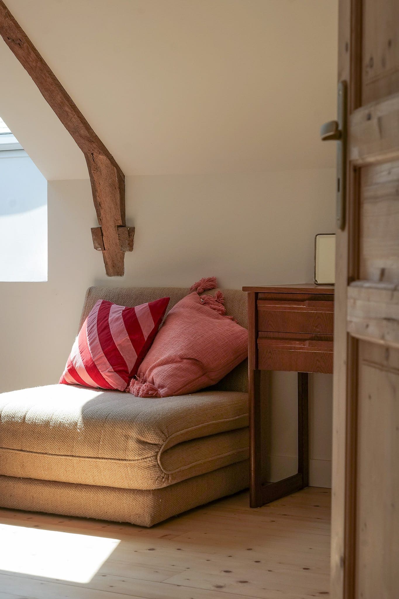 A cosy corner with a beige chair topped by two pink cushions, next to a wooden desk under a sloping ceiling with exposed beams and sunlight streaming through a nearby window.