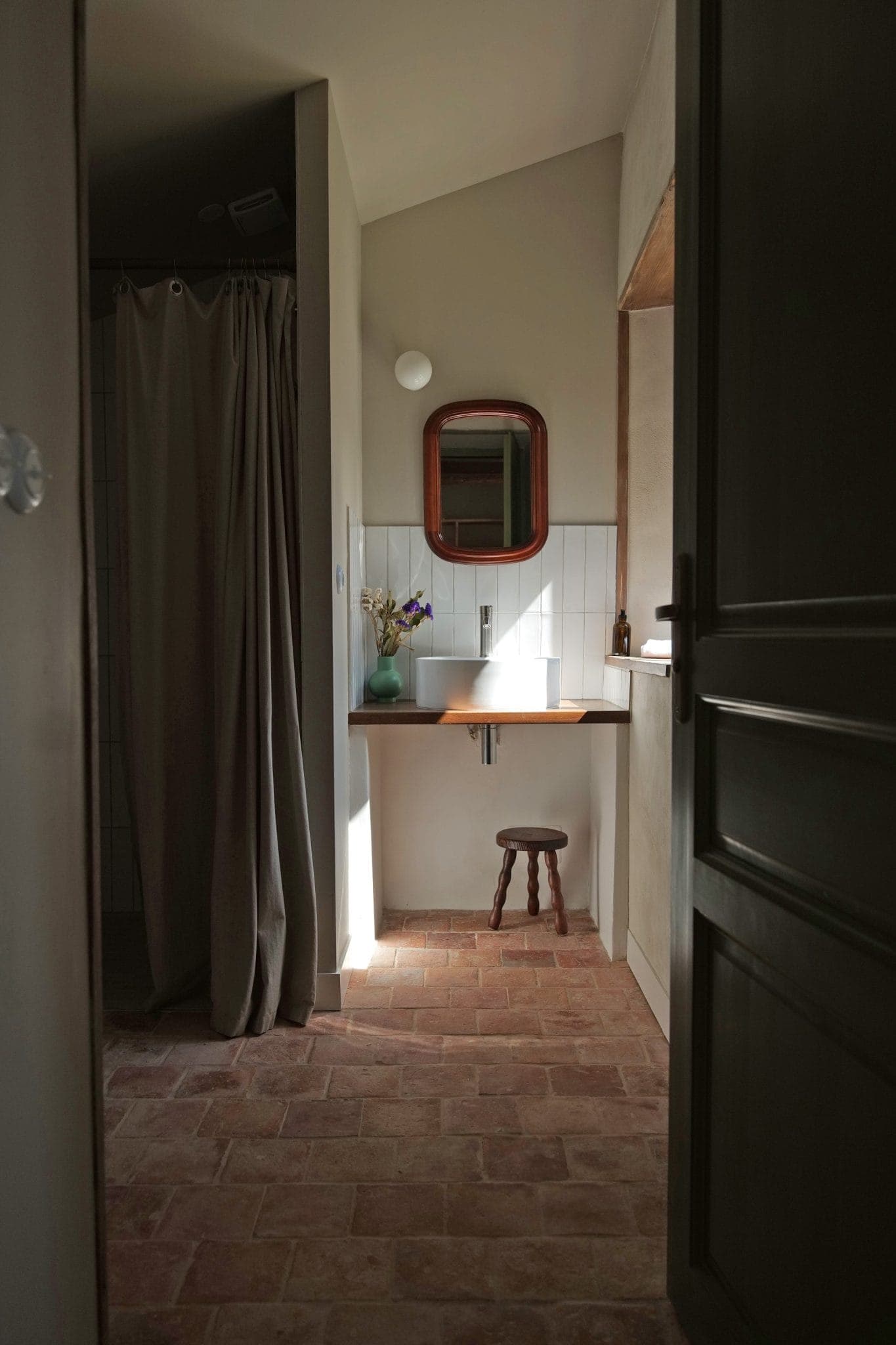 A softly lit bathroom with a tiled floor, a wooden stool under a small sink, an oval mirror above the sink, a vase with flowers and a shower area with a beige curtain on the left.