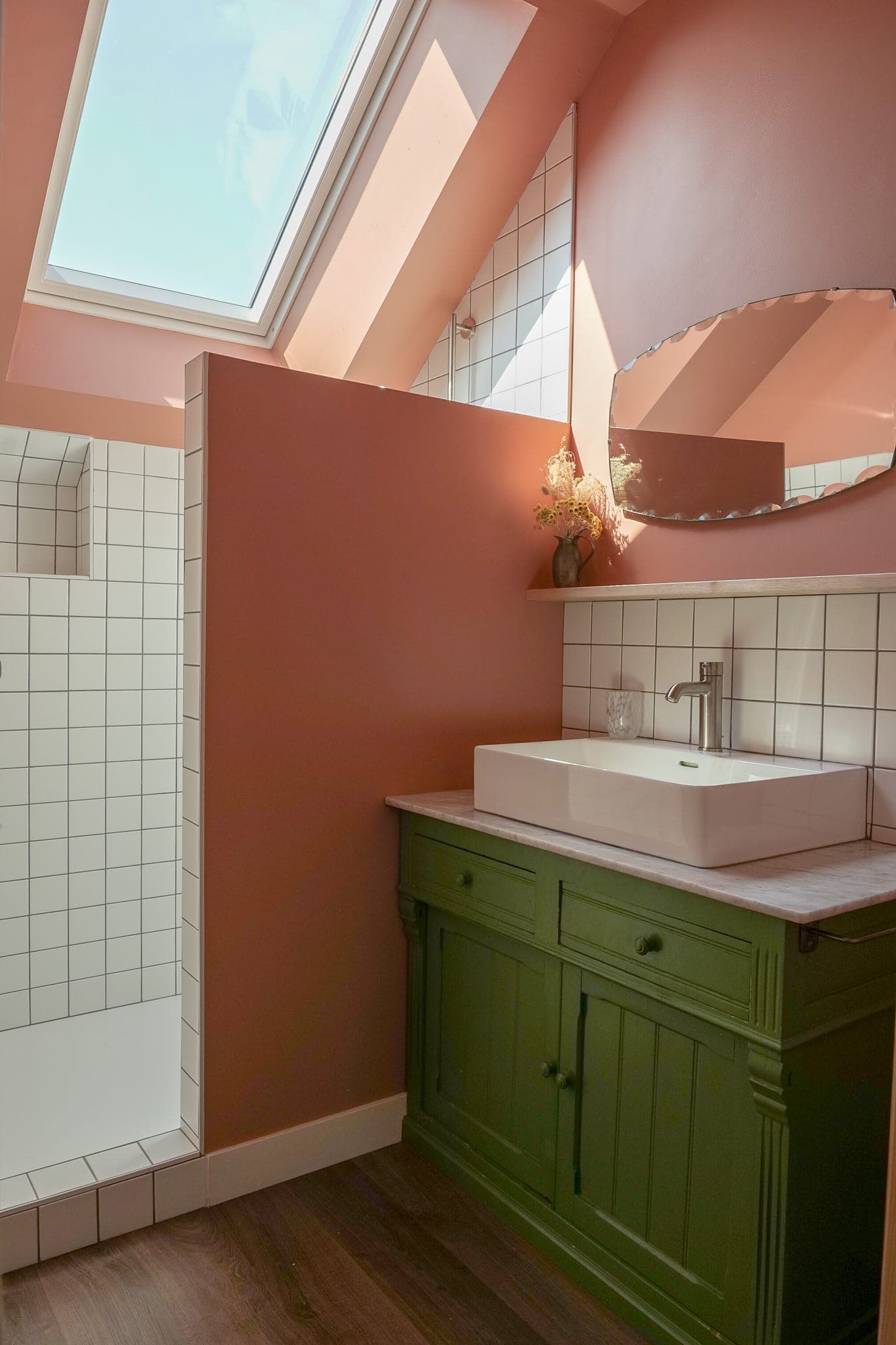 A bathroom with pink walls, a green vanity unit and a white sink, a round mirror, a white-tiled credenza and a white-tiled walk-in shower. A skylight lets in natural light.