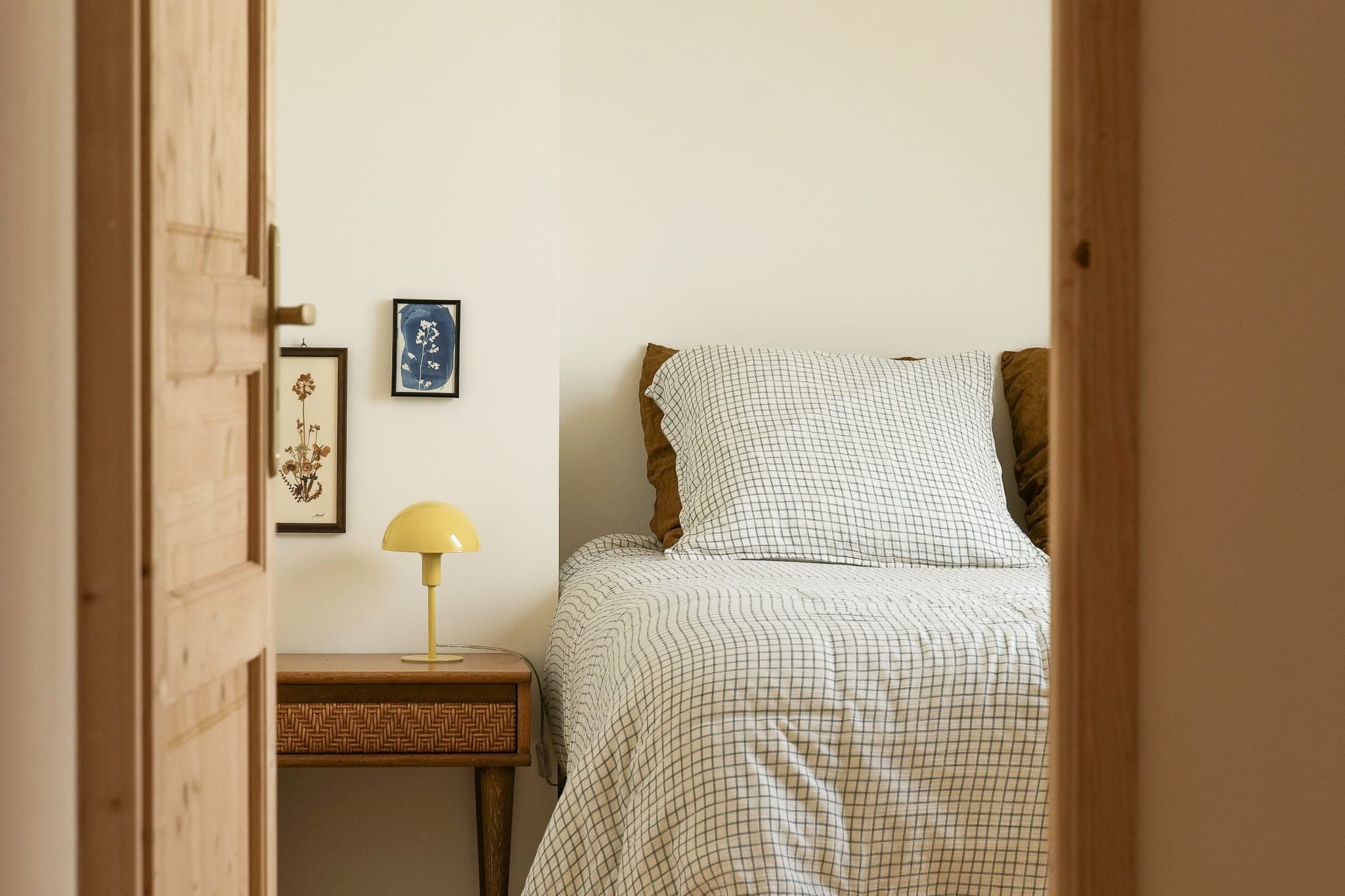 A comfortable bedroom with a neatly made bed with a checked comforter, a wooden bedside table with a small yellow lamp and two framed paintings on a cream-colored wall, seen through a partially open wooden door.