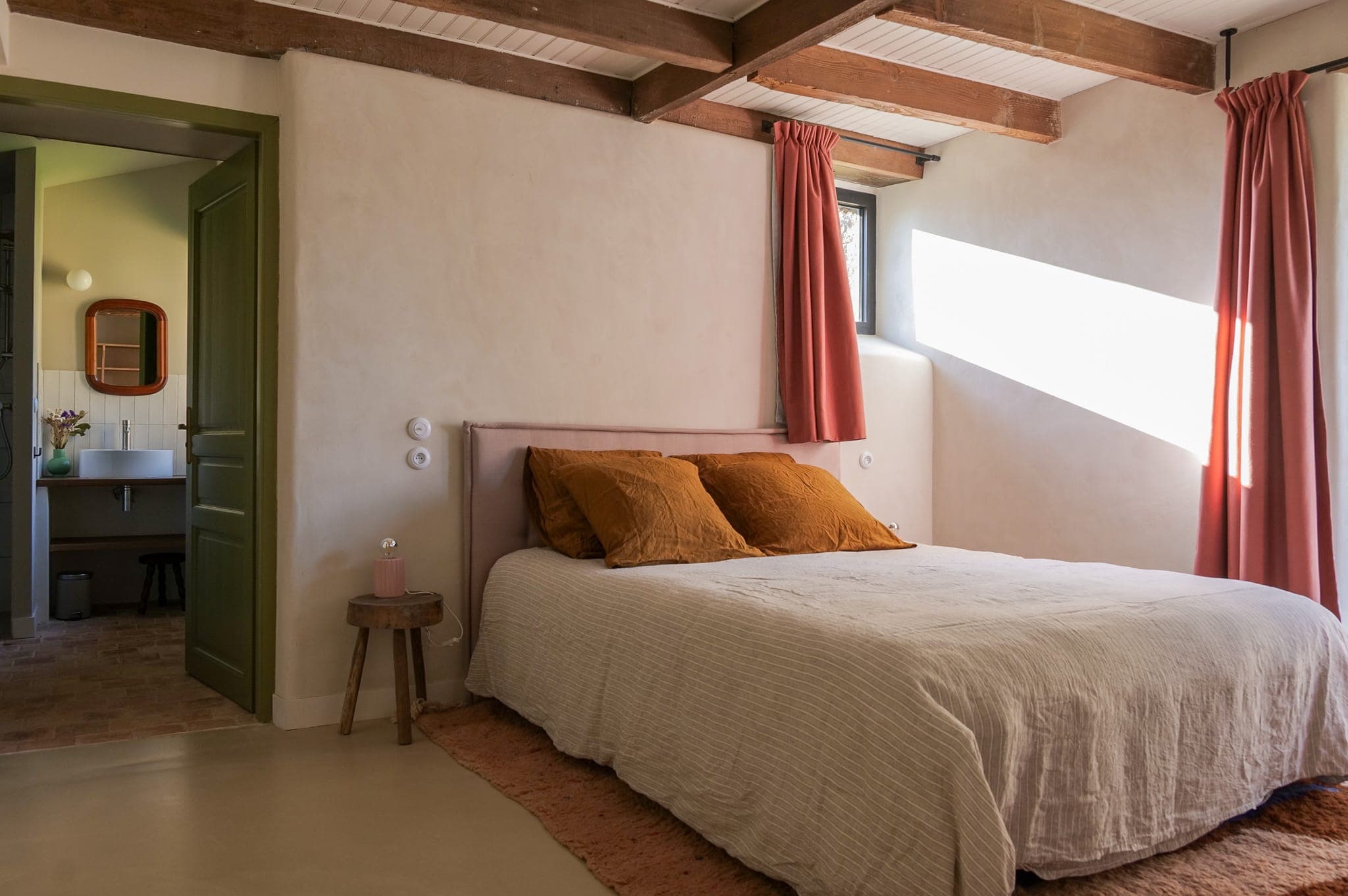 Bright room with double bed, mustard pillows, light bedding, pink curtains, small wooden side table and open door to bathroom with sink and mirror. Exposed ceiling beams.