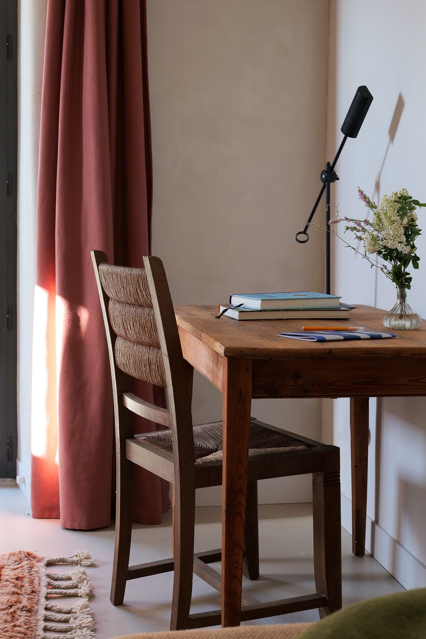 A wooden desk and chair sit next to a window with pink curtains. On the desk are books, a notebook, a pen, a modern black lamp and a small flowery vase. Sunlight floods into this warm, minimalist room.