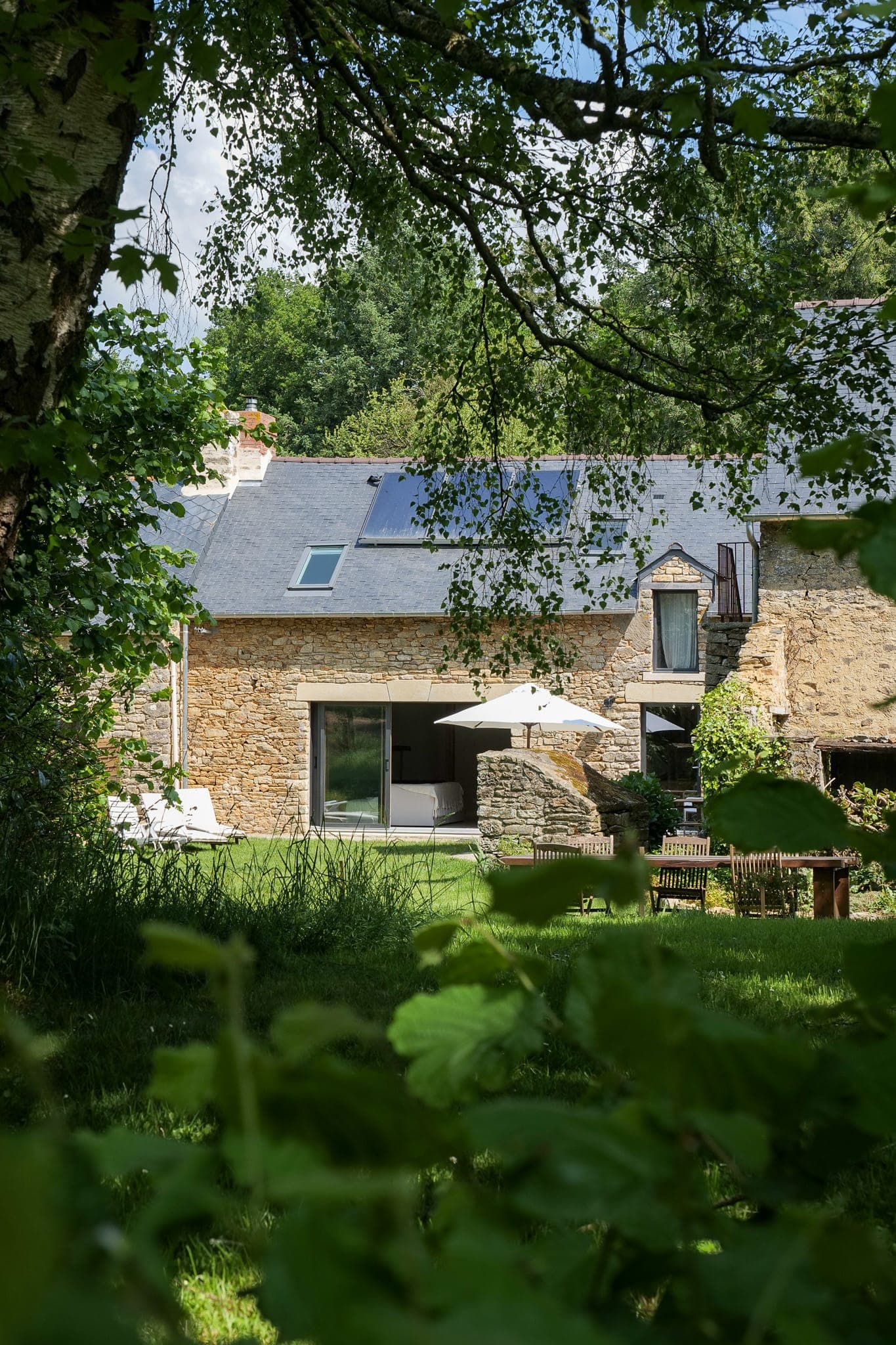 A rustic stone house with slate roof and solar panels is surrounded by greenery. A terrace with garden furniture and white parasol sits in the lush garden, bordered by trees and luxuriant plants.