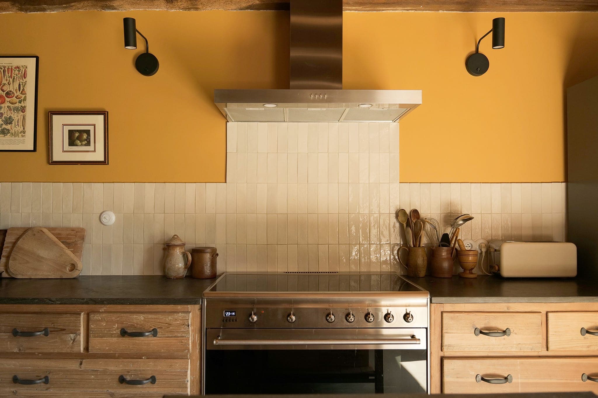 Une cuisine moderne avec des murs jaunes, des armoires en bois clair, un four et une hotte en acier inoxydable, des ustensiles de cuisine dans des bocaux, un grille-pain et des luminaires muraux au-dessus d'un dosseret carrelé.