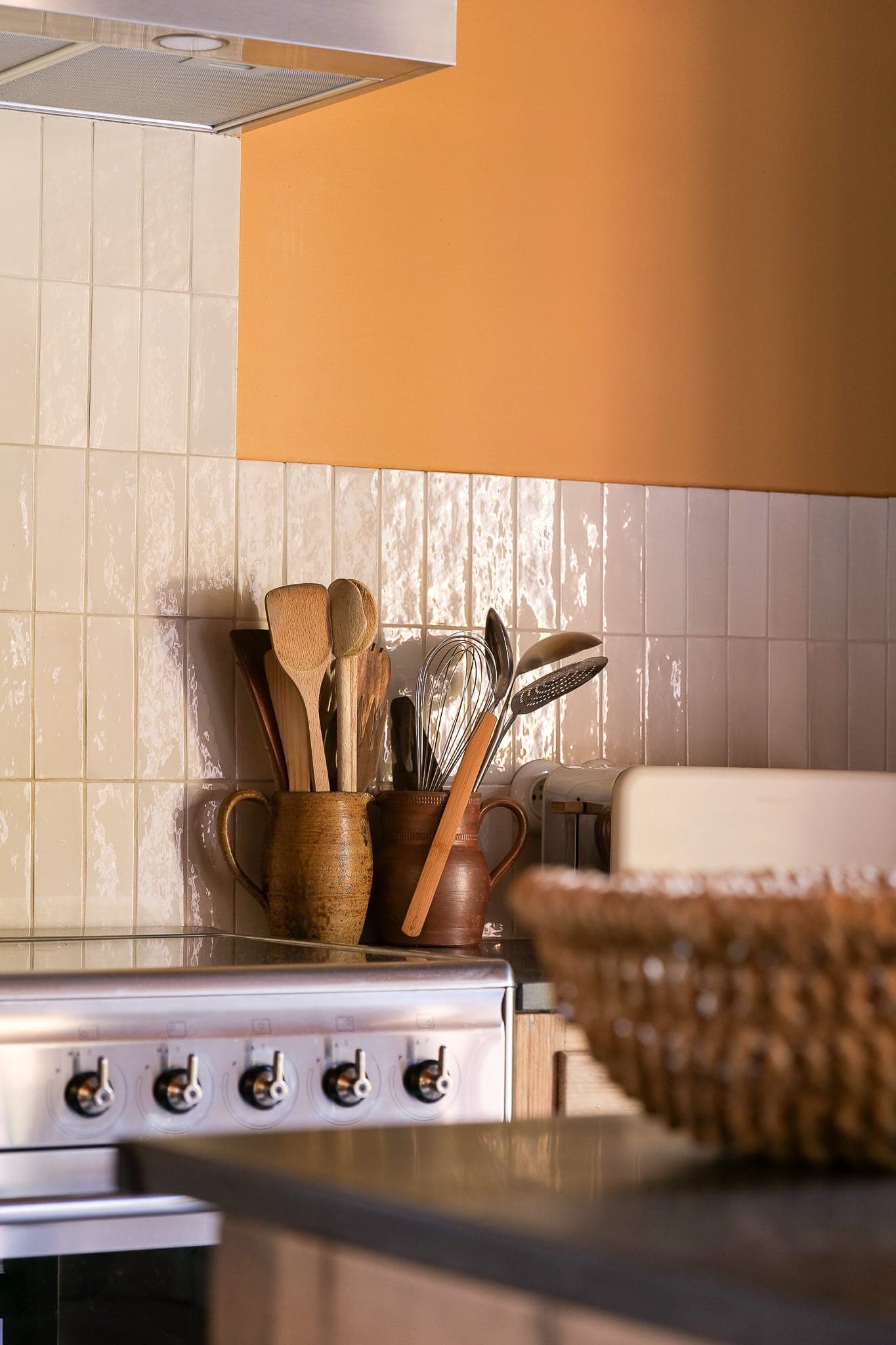 A kitchen worktop with wood and metal utensils in ceramic holders, next to a stove. The credenza is tiled in off-white, with a woven basket in the foreground. The wall is painted a warm beige.